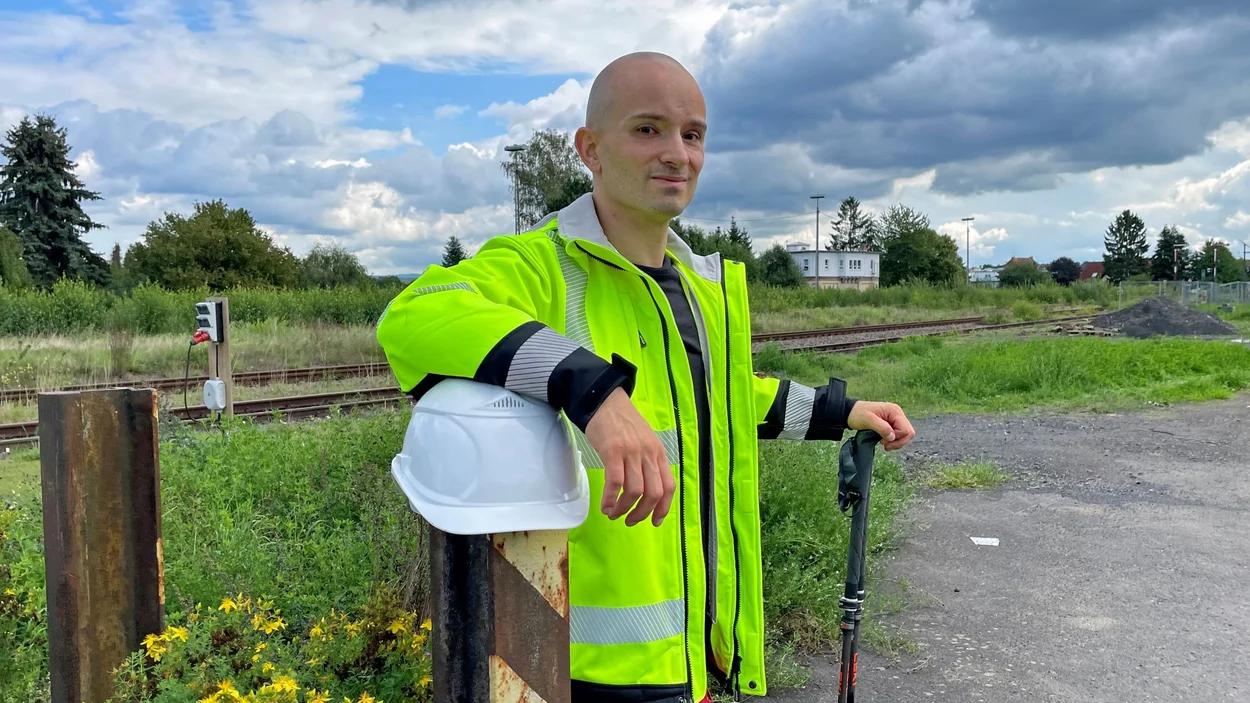 Nicolai Spatz lehnt mit leuchtend gelber Warnjacke und Stöcken an einem Pfosten, über den ein weißer Helm gestülpt ist