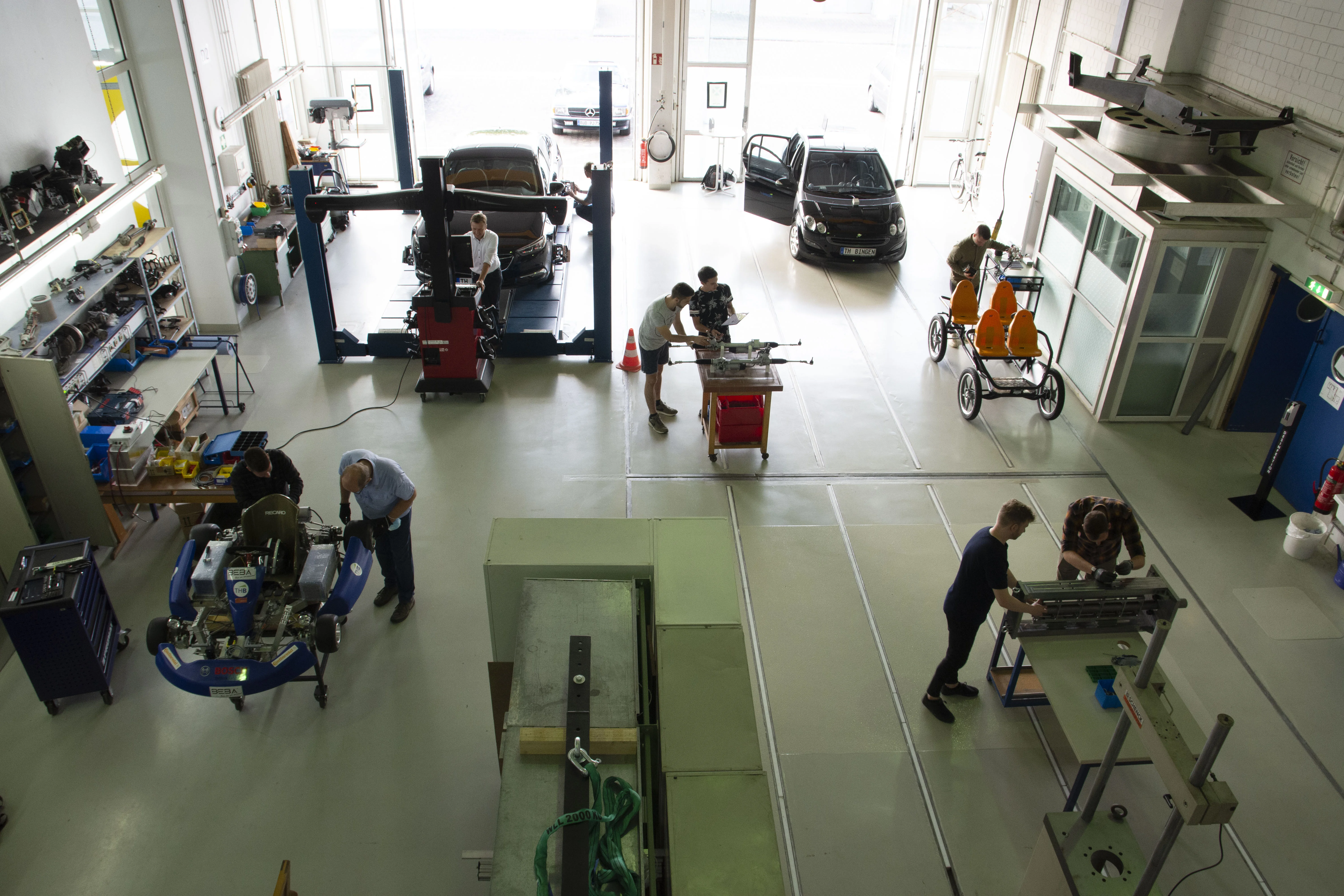 Blick von oben auf das Fahrzeuglabor der TH Bingen. Zu sehen sind einige Fahrzeuge und Fahrzeugteile sowie Personen, die an den Fahrzeugen und Fahrzeugteilen arbeiten.