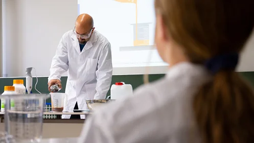 Auf dem Bild ist ein Professor im Labor in einem Laborkittel zu sehen, der eine Flüssigkeit in einen Messbecher leert.