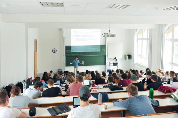 Das Bild zeigt Studierende in einem Hörsaal der Technischen Hochschule Bingen und einen Lehrenden vor einer großen Tafel, der gerade etwas erklärt.
