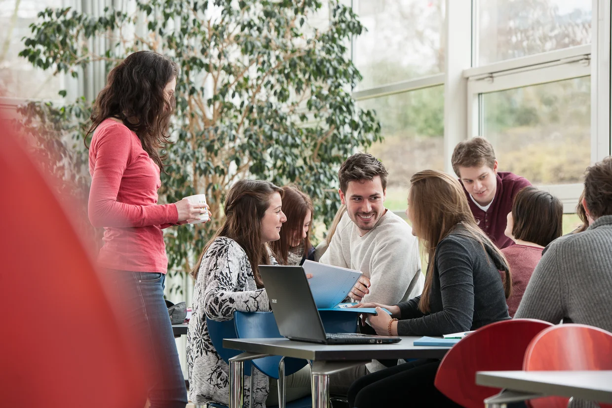 Studierende sitzen lachend am Tisch, eine Studentin mit Kaffeebecher steht vor dem Tisch