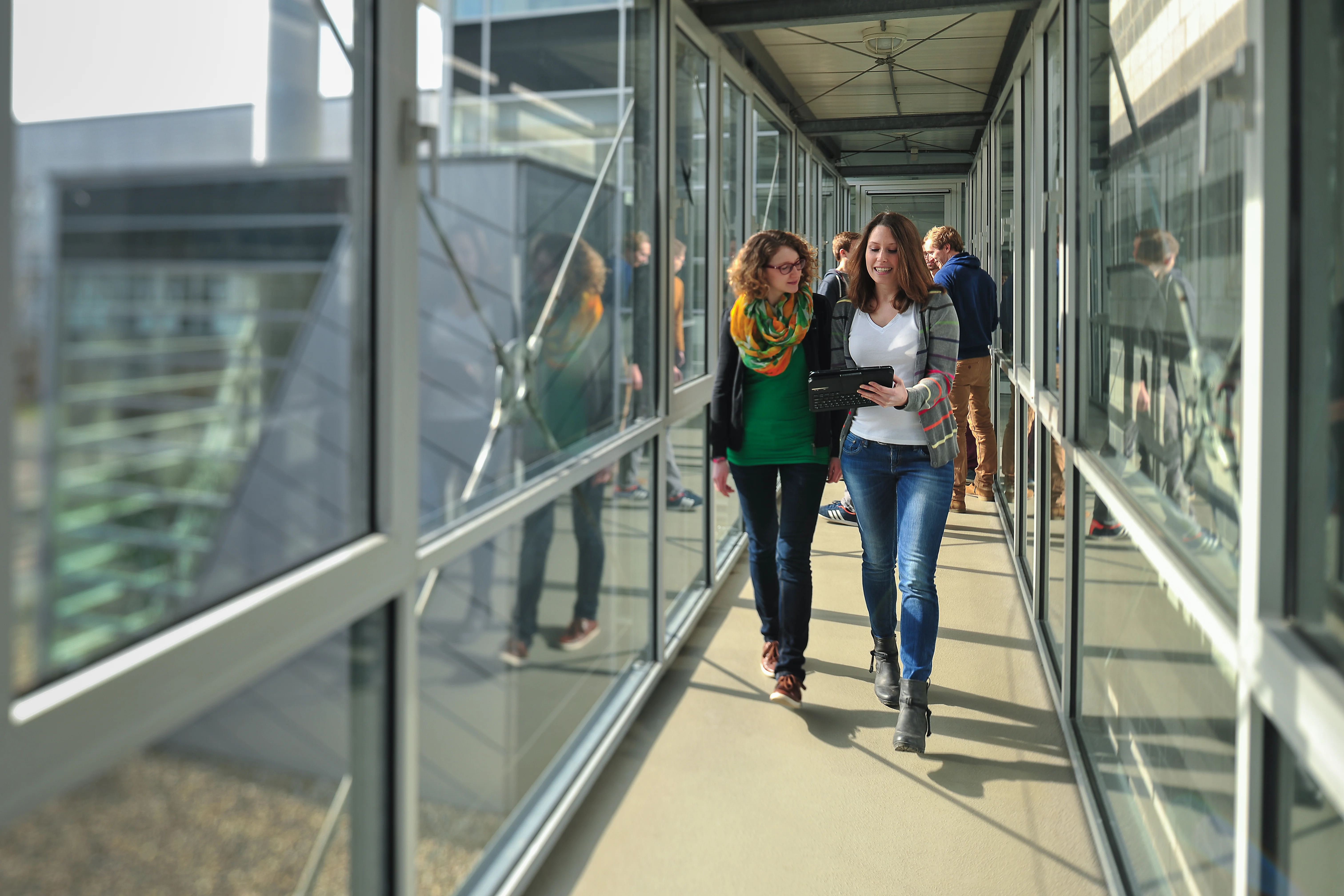 Das Bild zeigt zwei Frauen gemeinsam durch einen Flur der TH Bingen laufend. Eine trägt ein Tablet in der Hand. Die beiden unterhalten sich. 