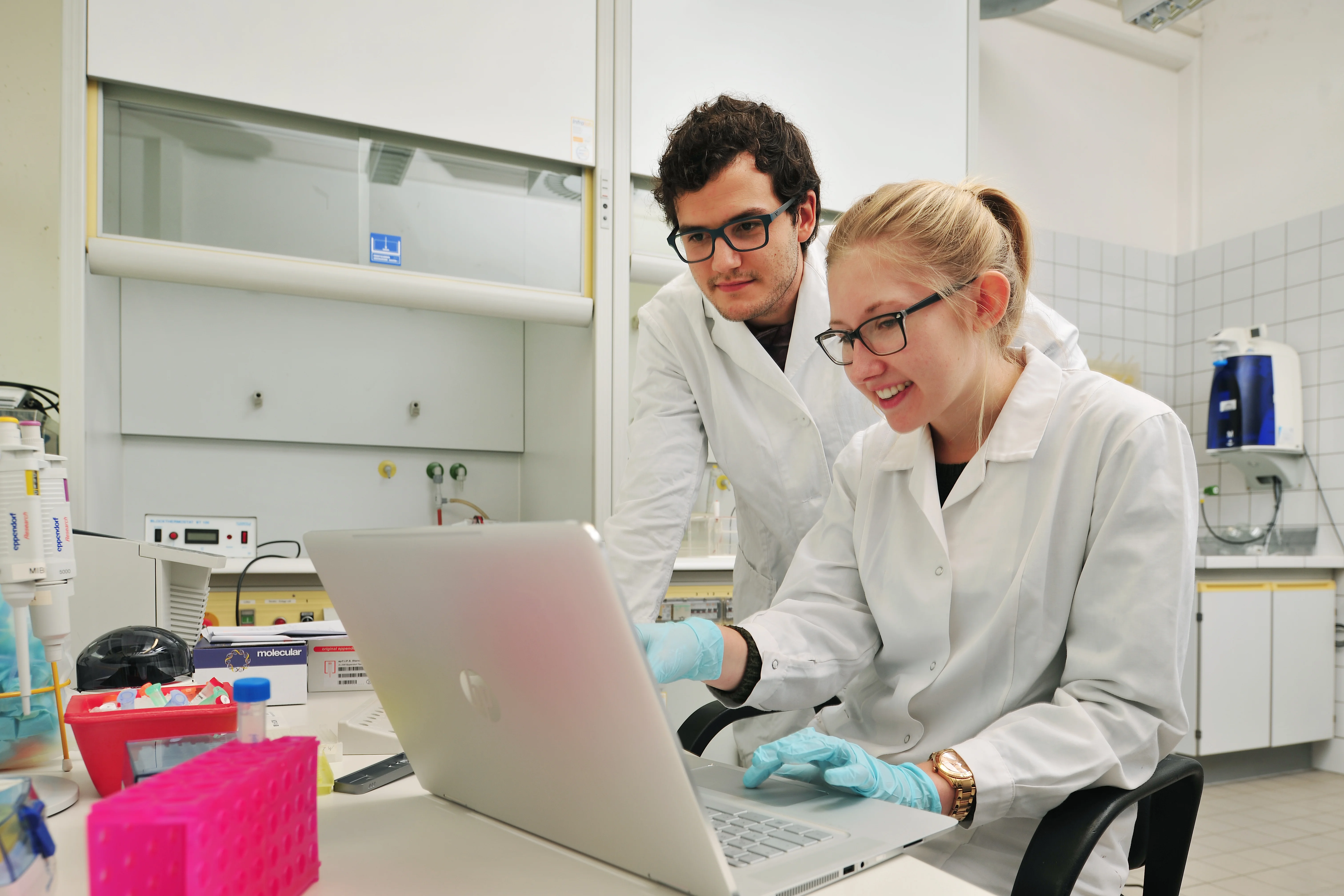 Das Bild zeigt zwei Studierende im Labor in Laborkittel und Schutzbrille vor einem Laptop.